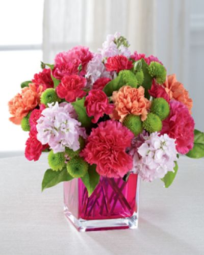 Dazzling and delightful, this bold and colorful flower bouquet is set to impress with it's high impact look and graceful styling. Hot pink carnations, orange carnations, pale pink gilly flower, hot pink mini carnations, green button poms, and lush greens are beautifully arranged in a raspberry pink glass cubed vase to create a rush of fun and warm wishes to send straight to your recipient's door.