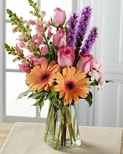Exuding a special charm, with a casual fresh-from-the-garden look, this gorgeous spring bouquet is the perfect way to delight your recipient in honor of any of life's most treasured moments. Peach gerbera daisies are soft and sophisticated surrounded by pink roses, pink snapdragons, pink mini carnations, purple liatris, and lush greens arranged with an artist's eye in a gathered square clear glass vase.